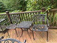 Two cast metal patio chairs and one matching side table on outdoor wooden deck next to railing and greenery.