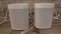 Front view of two white Sonos One speakers side by side on a floor surface, cables visible behind