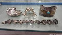 Overview of four jewelry pieces on display: swan and frog brooches, southwestern-style cuff bracelet, frog link bracelet