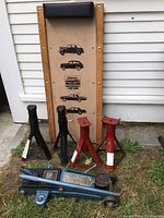Photo showing rolling car creeper with cushioned headrest, two black metal jacks, two red metal jack stands, and blue hydraulic floor jack placed on grass.