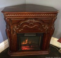 Front view of electric corner fireplace showing carved wood detail and glowing ember effect inside glass panel.