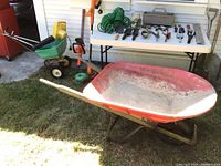 Overall outdoor photo showing red metal wheelbarrow with wooden handles, green hose, orange cultivator, yellow and green spreader, and white table with various hand tools spread out.