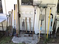 Photo showing the full lot of long handled garden tools including shovels, rakes, pitch fork, axes, and additional handles.