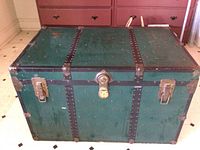 Front view of forest green metal trunk showing latch, lock, handles, and black riveted metal strips.