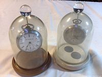 Both glass domes with Elgin watch on wooden base at left and Tivoli watch on white base at right.