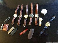 Top down image showing 9 wristwatches, 1 pocket watch, and various folding knives with leather cases on dark surface