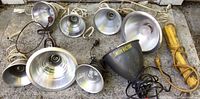 Eight clip-on work lights laid out on the ground. Various sizes, all with metal reflector shades and electric cords. One branded Smith Victor light in black. All lights visible with clip-on mounts.