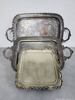 Three silver plated trays stacked, showing varying sizes and decorative handles. Tarnished surface visible.