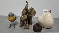 Front view of 4 animal display items including elephant with baby, ceramic chicken, wooden bird, and small brown bird on a gray surface.