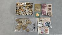 Photo of a metal tin filled with assorted foreign coins, a plastic bag with coins labeled 'MEX', and various foreign paper bills laid out.