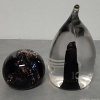 Both glass paperweights side by side on a neutral surface: round signed sphere with decorative elements and larger clear teardrop Kolter Waterclub paperweight