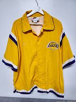 Front view of yellow Los Angeles Lakers Nike warmup shirt jacket showing Lakers logo and Nike swoosh on chest with short sleeves and stripe accents.