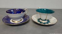 Photo showing two cup and saucer sets side by side, one Royal Albert with blue and gold design, and one Paragon with teal and white with gold accents.
