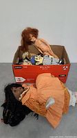 Two vintage dolls, one laying down naked with red hair, another dark-skinned doll in orange dress with black braided hair beside a shoebox with other dolls inside.