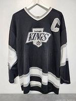 Front view of black CCM Los Angeles Kings hockey jersey with white and grey stripes, featuring the team logo and a captain's 'C' patch.