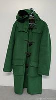 Front view of the green vintage wool jacket with hood and toggle fasteners visible, hanging on a hanger against a plain wall.