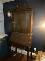 Full view of antique wooden secretary desk showing its overall shape, glass door cabinet, drop-front desk, and lower drawer.