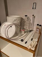 Plastic buckets with lids, clear tubing, glass carboy, hydrometer, and bottles of additives visible