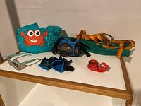 Photo showing puddle jumper floatation device, portable swing, protective gloves, blue and black elbow/knee guards, and red bike bell arranged on a white tabletop.