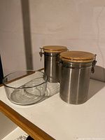 Clear photo showing two stainless steel lock-top containers with bamboo lids and one large glass bowl on a wooden shelf.