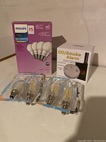 Box of Philips 60W LED bulbs, four individual LED filament bulbs in plastic packaging, and CO/Smoke Alarm box