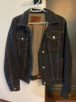 Dark blue denim jean jacket hanging open on white hanger, showing front design and interior lining.