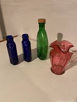 Photo showing two blue glass bottles, one green glass bottle with cork, and one cranberry glass pitcher on a white surface against a cream background.