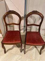Pair of wooden dining chairs with carved grape detail on curving backs, red upholstered seats, standing on floor against wall.