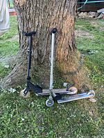 Two children's scooters leaning against a tree outdoors on grass, one black and one silver Razor scooter.
