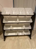 Front view of 4-tier storage shelf with 8 small and 4 large white plastic bins installed on metal rods in a dark wood frame.