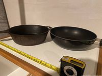 Two deep skillets placed side by side, with a tape measure in front for scale. The left skillet is cast iron with a small handle, and the right skillet is black with a plastic handle.