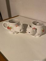 Two white kitten teacup holders with floral details placed on a flat surface.