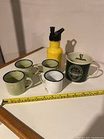 Photo of four enamelled metal cups (three green with blue rims, one white with blue rim) and one larger Guinness logo enamelled cup, along with a yellow child's water bottle with black spout, all displayed on a wooden surface with a tape measure.
