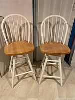 Two wooden bar stools with natural wood seats and white painted bases, showing spindle backs and swivel seats.
