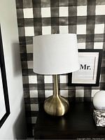 Photo of one side lamp with a brass or gold-toned rounded base and white fabric drum shade, placed on a dark wood surface against a black and white checkered wall