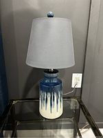 Photo of one of the two similar side lamps with ceramic base, blue and white drip glaze, and gray fabric shade placed on glass table.
