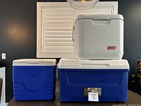 Three coolers set: one small white Coleman cooler on top of one large blue Igloo cooler with white lid, next to one medium blue Coleman cooler with white lid.