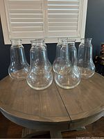 Seven large clear glass vases arranged on a wooden table, showing their size and shape.