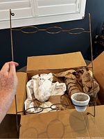 Photo of metal wall mount frame showing nine circular loops to hold pots, held above a cardboard box with wrapped items inside.