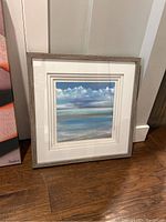 Framed art piece showing an ocean or sky view with clouds, placed on the floor.