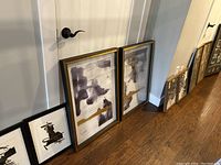 Photo shows several framed artworks leaned against the wall including two larger abstract paintings with gold accents and two smaller black and white pieces, some with visible water damage