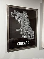 Framed typographic map of Chicago city showing neighborhood names in white text on black background with metal frame.