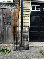 Old black metal wire dog cage leaning against a wooden fence and garage door outdoors, showing signs of wear and missing the plastic insert on the bottom.