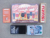 Top view of the lot showing Brew-opoly boxed game with beer glasses illustration, Mini-Jenga wooden block set, Cards Against Humanity black box, Dominos set box, and an orange tin labeled Magnetic Hangman.