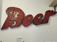 Large distressed red metal sign spelling the word 'Beer' in cursive style lettering, mounted on a wall, shows weathered paint effects.