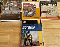 Four books: Concrete Manual, Foundations and Concrete Work, The Complete Guide to Plumbing, Fundamentals of Crew Leadership, displayed on a wooden table.