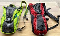 Two hydration backpacks by Camelbak, one green/gray and one red/black, laid flat to show front side with straps and buckles