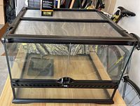Front view of rectangular glass terrarium with black metal frame and sliding doors, mesh ventilated canopy with light bulb on top visible.