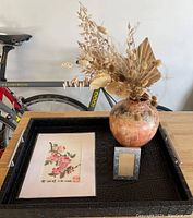 Photo showing black faux alligator skin serving tray holding framed floral print, small metal photo frame, and ceramic vase with dried flowers. Bicycle in background.
