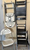 Four grey folding chairs stacked and five black folding chairs stacked beside them against a wall. Grey chairs have distinctive fan-shaped backrests and padded seats. Black chairs have a slatted seat and back design.